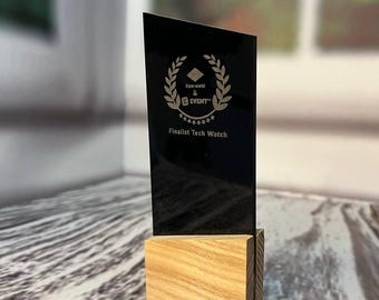 May include: Black acrylic award plaque with a gold laurel wreath and the text "Iron World & Event" and "Finalist Tech Watch". The plaque is mounted on a wooden base.
