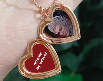 May include: A gold heart-shaped locket necklace with a red interior. The locket is open, revealing a photo of a man with white hair. The inscription on the locket reads "Astarion my beloved".