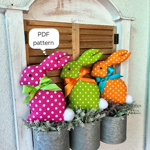 May include: Three fabric Easter bunnies in pink, green, and orange with white polka dots, each with a different coloured ribbon bow, sitting in galvanised metal planters with greenery.