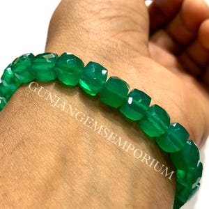 May include: A bracelet composed of faceted, vibrant green gemstone beads. The beads are uniform in size and shape, featuring a polished finish that reflects light. The text GUNJANGEMSEMPORIUM is overlaid across the center of the bracelet beads.