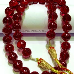 Puede incluir: Un collar de cuentas de piedras preciosas rojas con un borla dorada. Las cuentas son redondas y tienen un acabado brillante. La borla está hecha de hilo dorado y tiene un acento rojo.