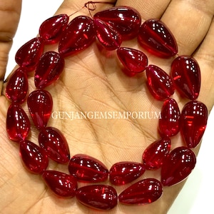May include: A strand of polished, teardrop-shaped, deep red gemstones. The gems are strung together, showcasing their translucent quality and rich color. The text "GUNJANGEMSEMPORIUM" is visible.