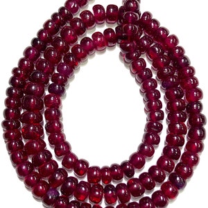 May include: A strand of deep red ruby beads, each bead is round and smooth, the beads are strung together to form a long necklace.