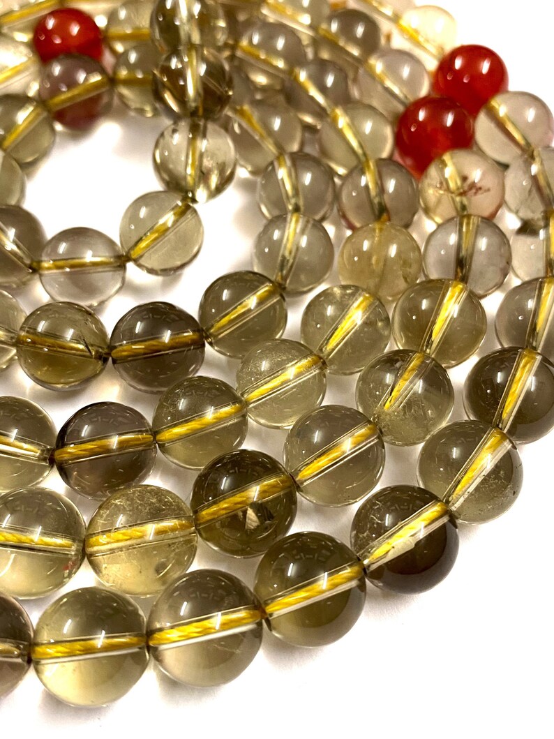 May include: A strand of smoky quartz beads with gold accents. The beads are round and translucent, with a light brown color. The gold accents are thin lines that run around the circumference of the beads.
