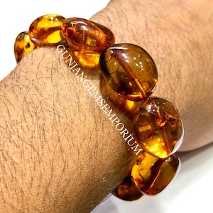 Könnte beinhalten: Ein Armband aus polierten, durchscheinenden, goldbraunen Bernsteinperlen mit unregelmäßigen, abgerundeten Formen. Die Perlen weisen natürliche Einschlüsse und Variationen in der Farbtiefe auf. Der Text GUNJANGEMSEMPORIUM ist über die Perlen gelegt.