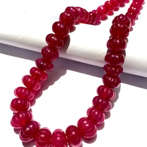 Puede incluir: Una hebra de cuentas de piedras preciosas de color rojo intenso con una superficie tallada y texturizada. Las cuentas están enhebradas en un hilo transparente y están dispuestas en un patrón circular.