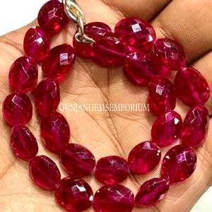 Puede incluir: Un collar de piedras preciosas rojas profundas, facetadas y de forma ovalada. Las piedras están ensartadas con un cierre plateado. El texto "GUNJANGEMSEMPORIUM" es visible. Las piedras se sostienen en una mano.