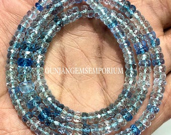 May include: Strands of faceted, light blue and clear gemstone beads. The beads are strung together, forming multiple loops. The image includes the text "GUNJANGEMSEMPORIUM". The beads are a mix of light and dark blue hues.