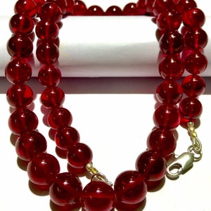以下が含まれることがあります： 大きな丸い半透明の赤いビーズのネックレス。ビーズは一緒に紐で繋がれており、二重のストランドを形成しています。銀色の留め金がネックレスを固定しています。ビーズは光沢のある仕上がりです。