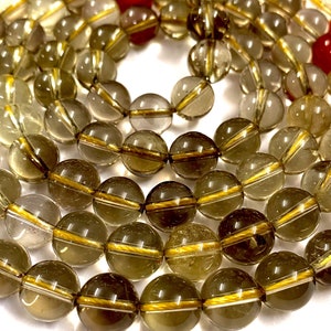 May include: A strand of smoky quartz beads with gold accents. The beads are round and translucent, with a light brown color. The gold accents are thin lines that run around the circumference of the beads.