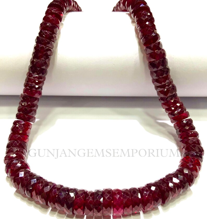 May include: A strand of deep red gemstone beads, likely ruby, with a faceted, barrel-shaped design. The beads are strung on a clear thread and are approximately 10mm in diameter.
