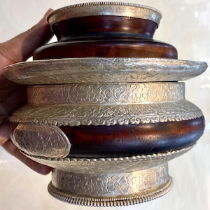 Puede incluir: Un recipiente decorativo antiguo con un cuerpo de madera marrón oscuro y adornos de metal plateado ornamentados. El recipiente presenta intrincados patrones florales y una pequeña placa grabada. El diseño sugiere un significado cultural o histórico.