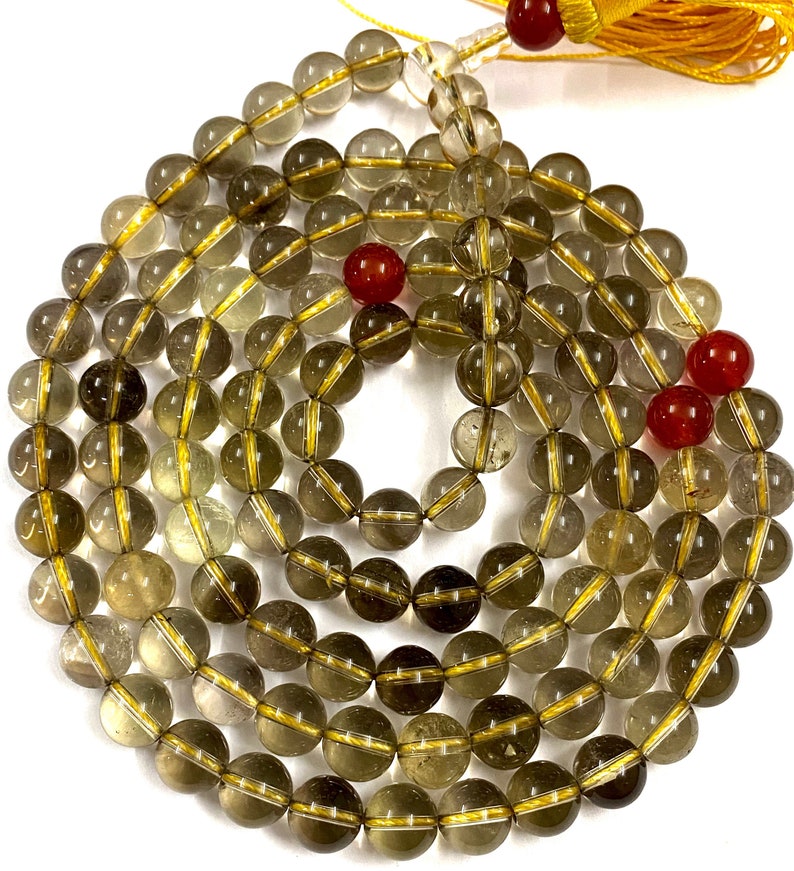 May include: A 108-bead mala necklace made of smoky quartz with gold accents. The beads are round and have a translucent appearance. The necklace is finished with a tassel.