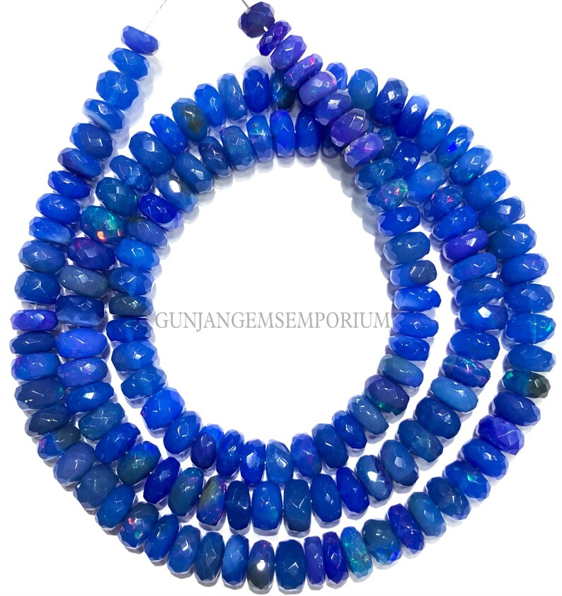 May include: A strand of blue opal beads with a faceted, barrel shape. The beads are strung on a clear thread and are approximately 10mm in diameter. The beads have a slight iridescent quality.