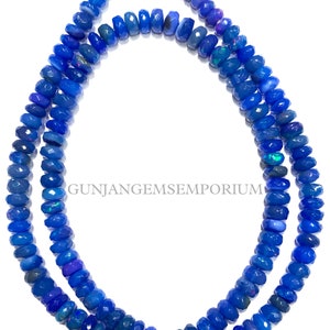 May include: A strand of blue opal beads, faceted and with a slight iridescent sheen. The beads are strung on a clear thread and are approximately 6mm in diameter. GUNJANGEMSEMPORIUM is printed on the white background.