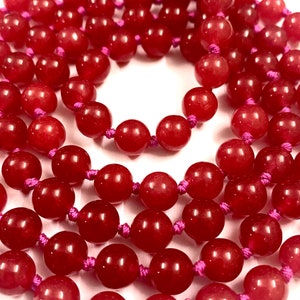 Könnte beinhalten: Nahaufnahme einer Halskette aus runden, tiefroten Perlen. Die Perlen sind mit einer leuchtend rosa Kordel aufgefädelt, wodurch ein geschichteter Effekt entsteht. Die Perlen haben eine glänzende Oberfläche und sind gleichmäßig angeordnet.