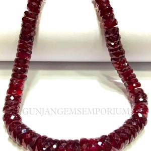 May include: A strand of deep red gemstone beads, each bead is faceted and has a cylindrical shape. The beads are strung together to form a necklace.