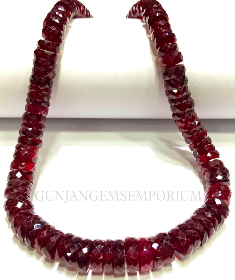 May include: A strand of deep red gemstone beads, each bead is faceted and has a cylindrical shape. The beads are strung together to form a necklace.
