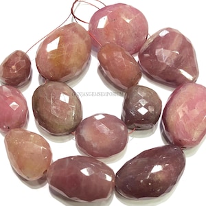 May include: A strand of faceted, irregular pink and mauve gemstone beads with a polished finish. The beads are strung on a thin red cord, and the text GUNJANGEMPORIUM is printed in small black letters near the center of the arrangement.