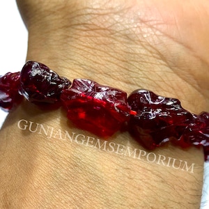 Peut inclure: Un bracelet composé de plusieurs perles de pierres précieuses rouge foncé, translucides, de forme irrégulière et facettées, enfilées sur un cordon rouge fin. Le texte GUNJANGEMSEMPORIUM est superposé au centre des perles du bracelet.
