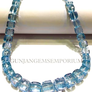 May include: A strand of light blue gemstone beads, each bead is faceted and has a square shape. The beads are strung on a clear thread and are arranged in a circular pattern. The beads are all approximately the same size and are evenly spaced. The image is taken from a close-up perspective, and the beads are in focus. The background is a white surface.