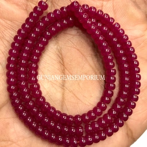 Puede incluir: Múltiples hebras de cuentas de piedras preciosas pulidas de color rojo intenso. Las cuentas son redondas y de tamaño uniforme, ensartadas juntas. La imagen muestra las cuentas en una mano, con el texto "GUNJANGEMSEMPORIUM" visible.