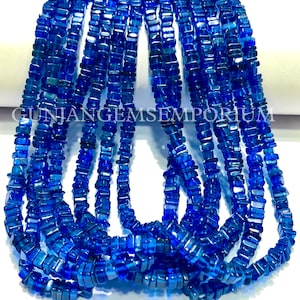 Puede incluir: Una hebra de cuentas de piedras preciosas azules, cada cuenta tiene forma cuadrada con un corte ligeramente irregular. Las cuentas están enhebradas en un hilo transparente. Las cuentas miden aproximadamente 10 mm de largo y 6 mm de ancho. GUNJANGEMSEMPORIUM está impreso en una superficie blanca detrás de las cuentas.