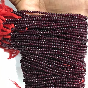 Può includere: Un filo di perline di pietra preziosa sfaccettata rosso scuro. Le perline sono piccole e uniformi per dimensioni e forma. Il filo è avvolto attorno a una mano.