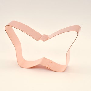 May include: A copper-coloured butterfly-shaped cookie cutter. The cutter is made of a single piece of metal bent into the shape of a butterfly. The wings are open, and the body is slightly curved. The cutter is designed for baking.