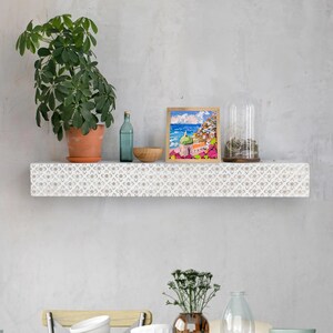 May include: White shelf with a carved floral pattern, featuring a framed painting of a coastal town, a glass cloche with greenery, and a clear glass bottle.