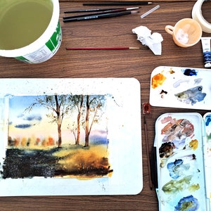 May include: A watercolor painting of a forest scene with trees and a sunset sky. The painting is on a white mat board and is surrounded by art supplies, including paintbrushes, paint tubes, a palette, and a cup of water.
