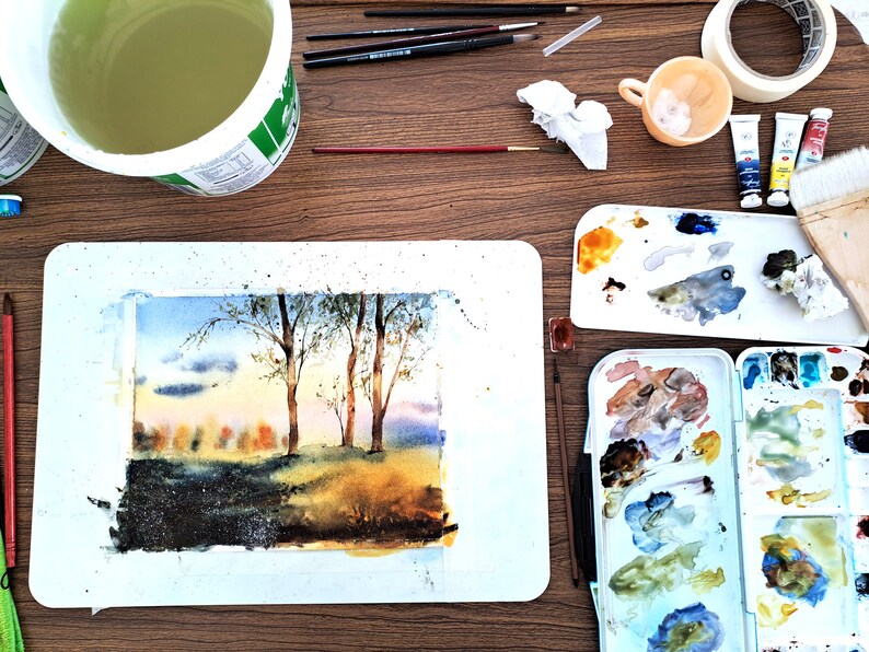 May include: A watercolor painting of a forest scene with trees and a sunset sky. The painting is on a white mat board and is surrounded by art supplies, including paintbrushes, paint tubes, a palette, and a cup of water.