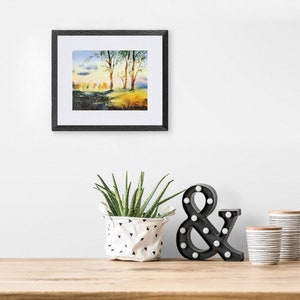May include: A framed watercolor painting of a forest scene with trees and a blue sky. The painting is hanging on a white wall above a wooden table with a potted plant, a black ampersand sign, and two white and black striped cups.