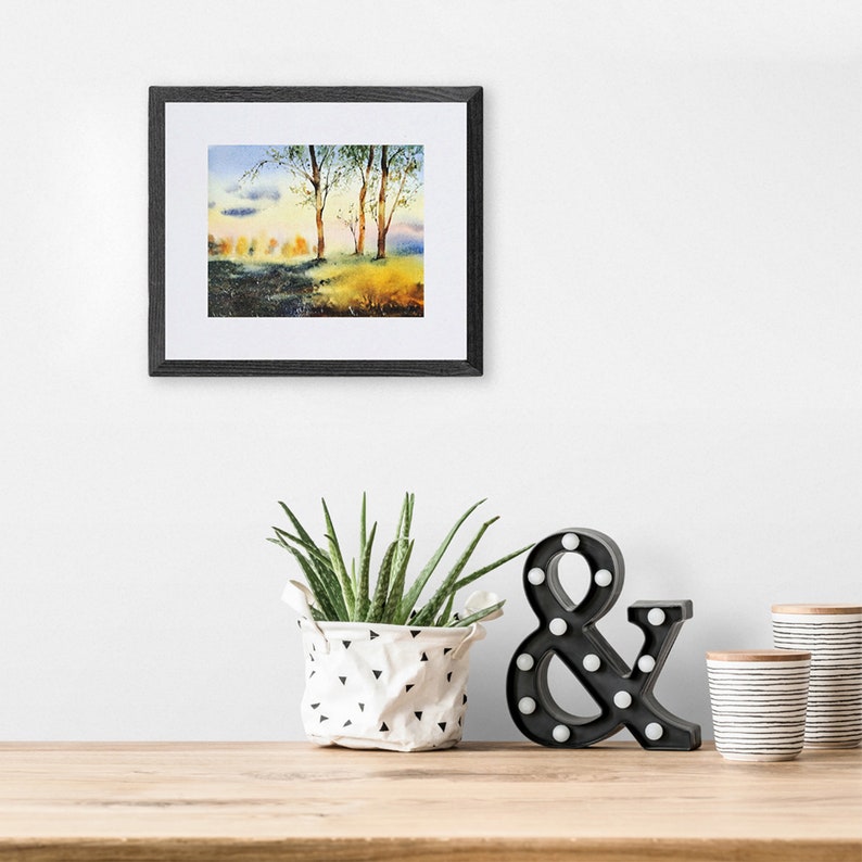 May include: A framed watercolor painting of a forest scene with trees and a blue sky. The painting is hanging on a white wall above a wooden table with a potted plant, a black ampersand sign, and two white and black striped cups.