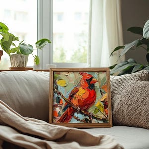 May include: A framed painting of a red cardinal perched on a branch with green leaves. The painting is in a wooden frame and is sitting on a white couch with a brown throw blanket.