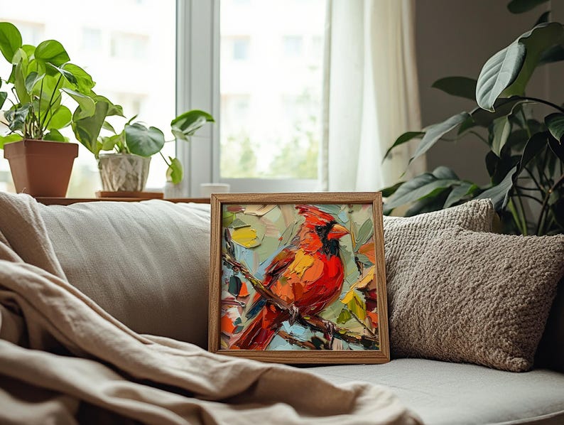 May include: A framed painting of a red cardinal perched on a branch with green leaves. The painting is in a wooden frame and is sitting on a white couch with a brown throw blanket.