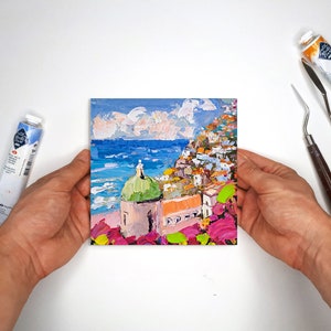 May include: A colorful painting of a seaside town with a green-domed building in the foreground. The town is built on a hillside and the sea is blue with white waves. The sky is a light blue with white clouds.