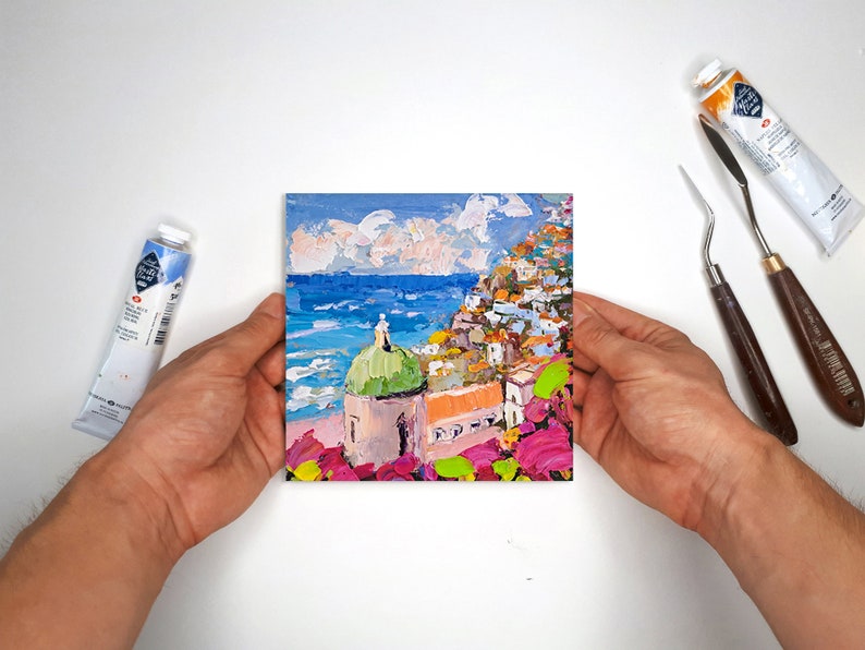 May include: A colorful painting of a seaside town with a green-domed building in the foreground. The town is built on a hillside and the sea is blue with white waves. The sky is a light blue with white clouds.