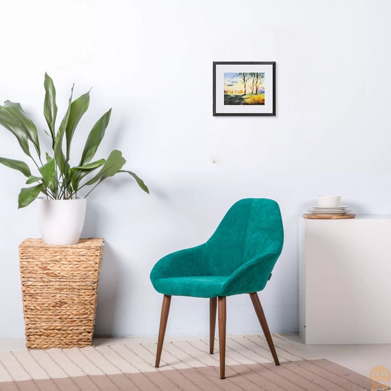 May include: A teal blue velvet armchair with wooden legs. The chair is in front of a white wall with a framed landscape painting above it.