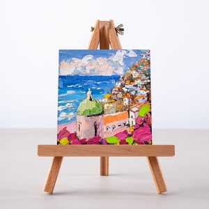 May include: A small painting on a wooden easel depicts a colorful Italian village with a green-domed church, overlooking a blue sea with white waves. The foreground features pink flowers.