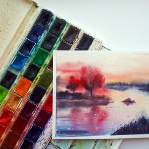May include: Watercolor painting of a landscape with a river and trees. The artwork features a vibrant sunset with red and orange hues. A palette of watercolor paints is visible in the background.