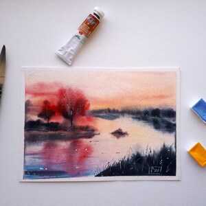 May include: Watercolor painting of a landscape with a lake and trees. The artwork features a vibrant sunset with red and orange hues. A paintbrush, a tube of paint, and two paint pans are next to the painting.