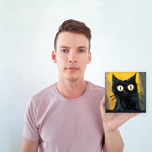 May include: A person holding a small painting of a black cat with large yellow eyes against a yellow background.