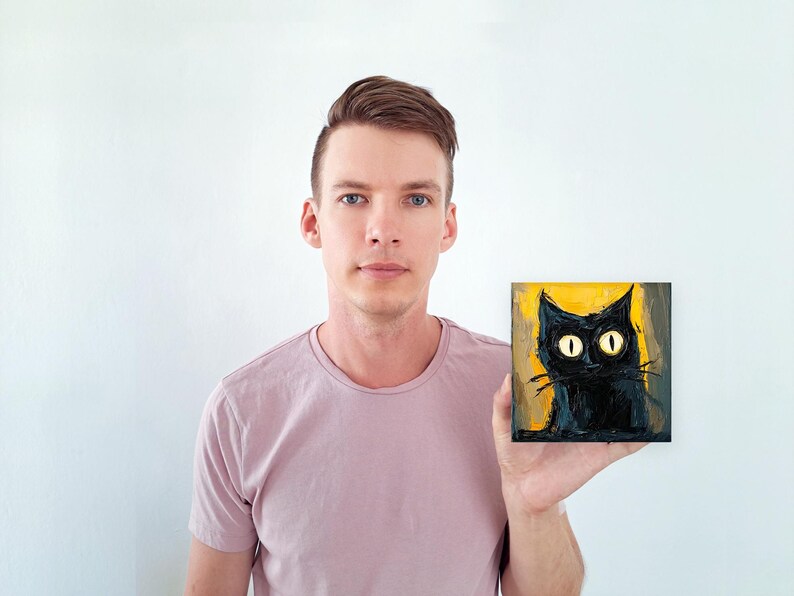 May include: A person holding a small painting of a black cat with large yellow eyes against a yellow background.