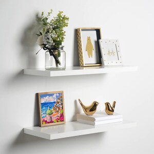 May include: Two white floating shelves with decorative items. The top shelf has a vase with white flowers, a gold framed picture of a bear, and a gold framed picture with two birds. The bottom shelf has a gold framed picture of a cityscape, two gold bird figurines, and a stack of books.