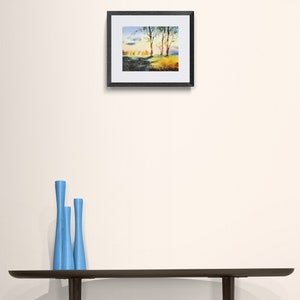 May include: A framed watercolor painting of a landscape with trees and a blue sky. The painting is in a black frame and is hanging on a white wall.