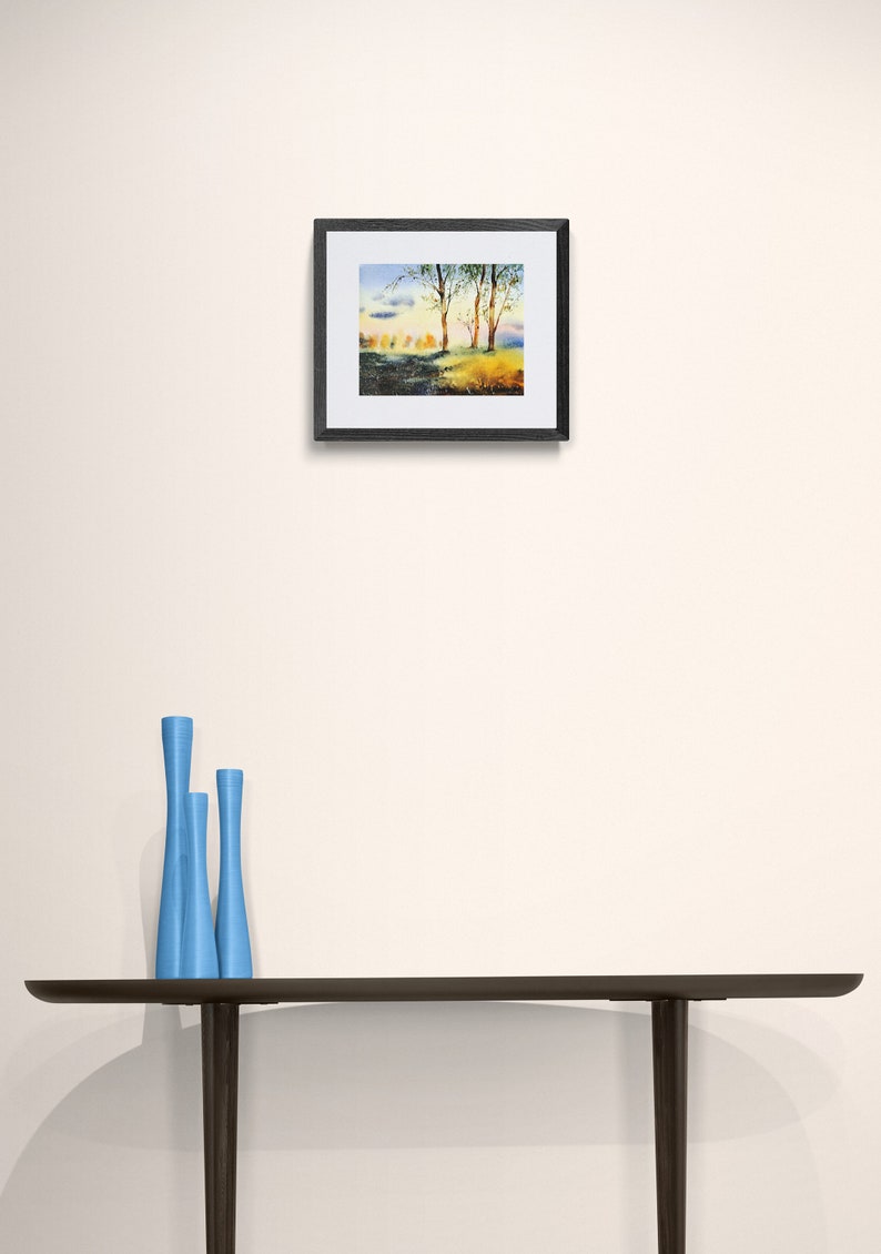 May include: A framed watercolor painting of a landscape with trees and a blue sky. The painting is in a black frame and is hanging on a white wall.