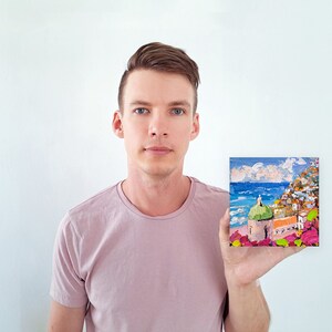 May include: A person holding a small painting of a colorful seaside town with a green domed building. The painting is set against a blue sky with white clouds.