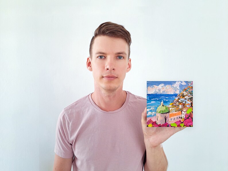May include: A person holding a small painting of a colorful seaside town with a green domed building. The painting is set against a blue sky with white clouds.