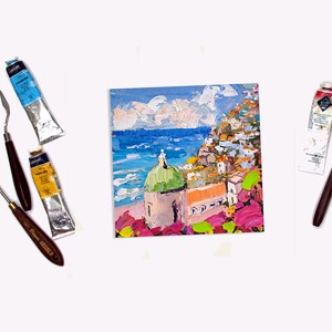 May include: A painting of a colorful Italian village with a green domed church, overlooking a blue ocean. The painting is done in a textured style with visible brushstrokes. The painting is surrounded by paint tubes and palette knives.